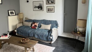 Ein Wohnzimmer mit einem blau gemusterten Sofa, einem kleinen Holztisch, einem Korbtisch mit einer Lampe und einem gerahmten Bild an der Wand.