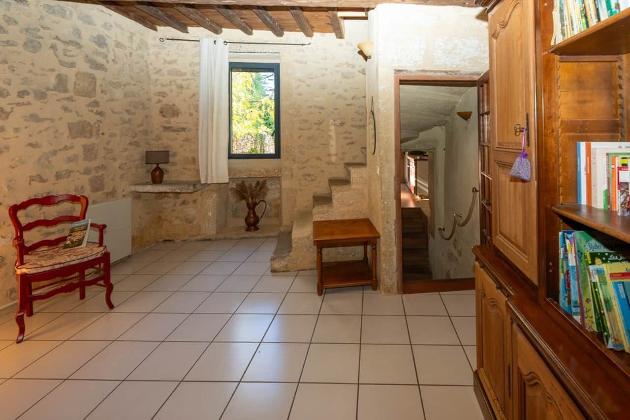 Une pièce rustique aux murs de pierre, dotée d’un sol carrelé, d’un banc en bois, d’une bibliothèque et d’une fenêtre donnant sur un paysage verdoyant.