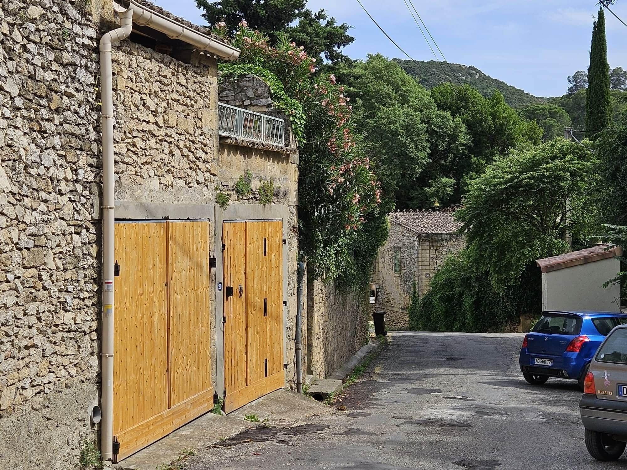 Un bâtiment en pierre avec des portes en bois, une rue étroite, des voitures garées et une végétation luxuriante.