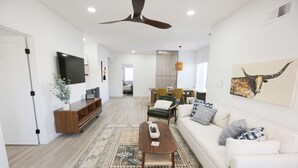 A spacious living room with a large TV, a ceiling fan, a patterned rug, a sofa, and a dining area with chairs. - Mission Gorgeous - Complete Remodel (Scottsdale)