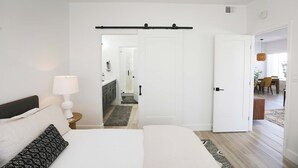 A bedroom with a bed, bedside table, lamp, and a door leading to a bathroom and dining area. - Mission Gorgeous - Complete Remodel (Scottsdale)