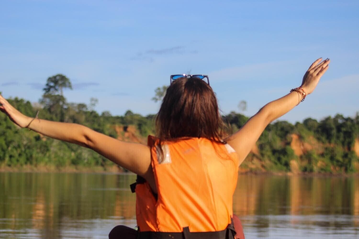 Una persona con una camisa naranja, con los brazos extendidos, mirando un cuerpo de agua y árboles al fondo.
