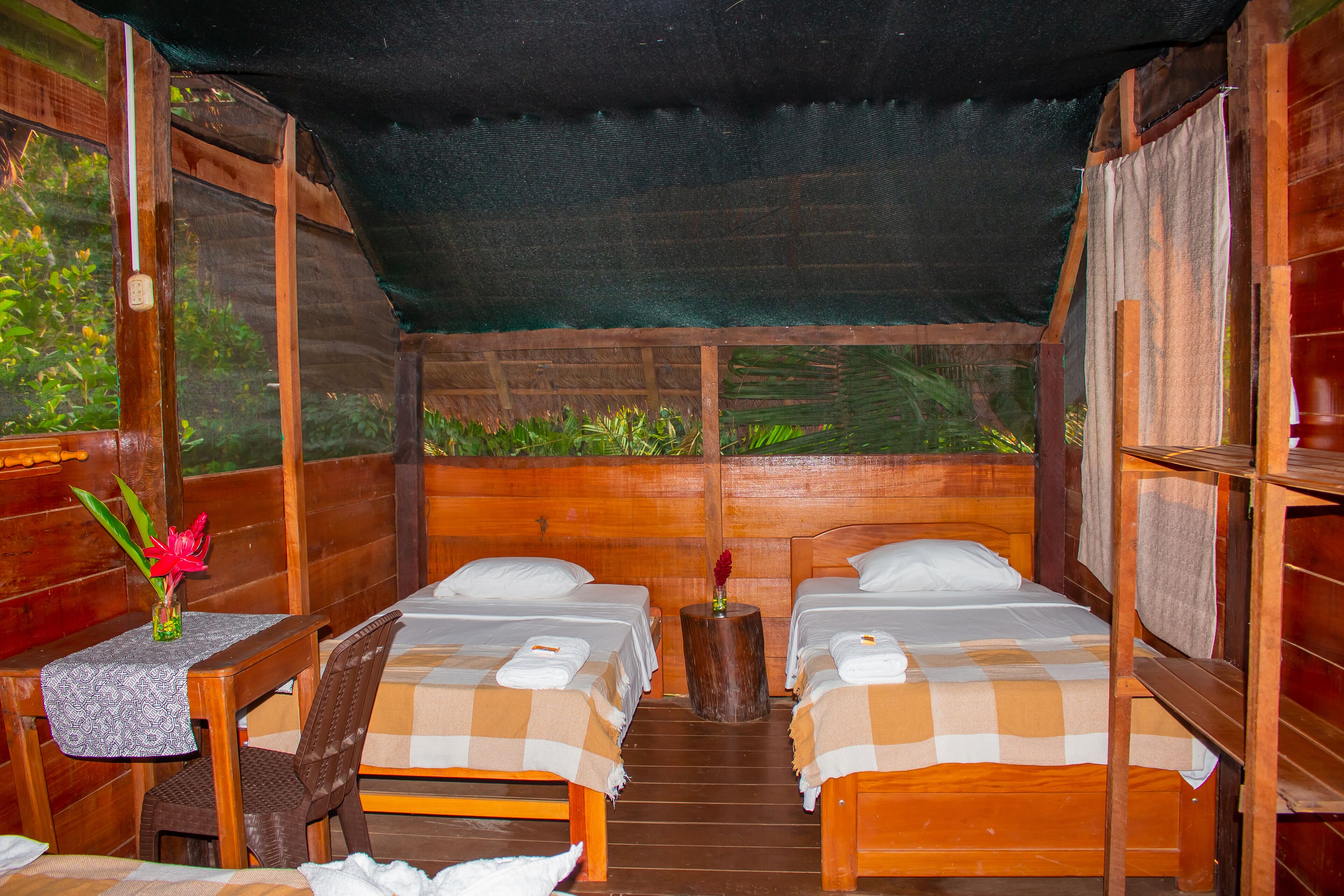 A wooden cabin with two beds, a small table with a vase, and a view of greenery outside.
