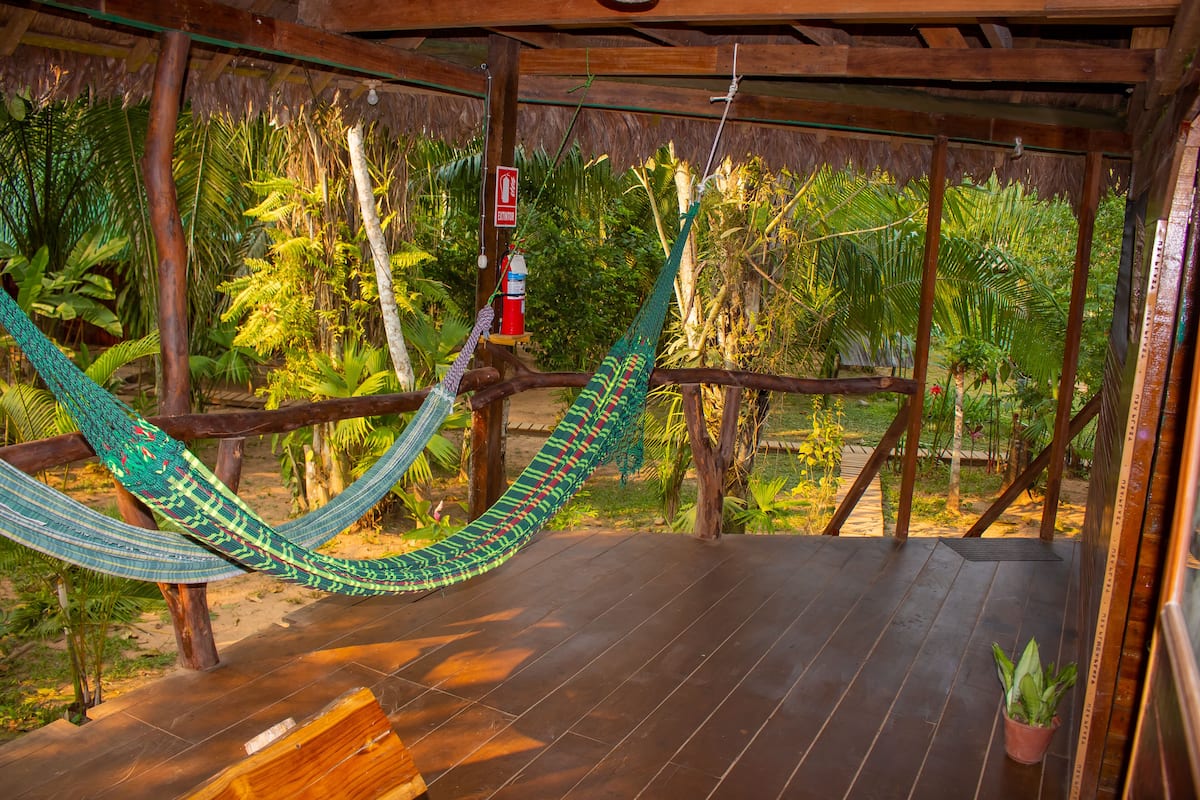 A hammock suspended between wooden beams in a covered outdoor area with a wooden deck and tropical plants.