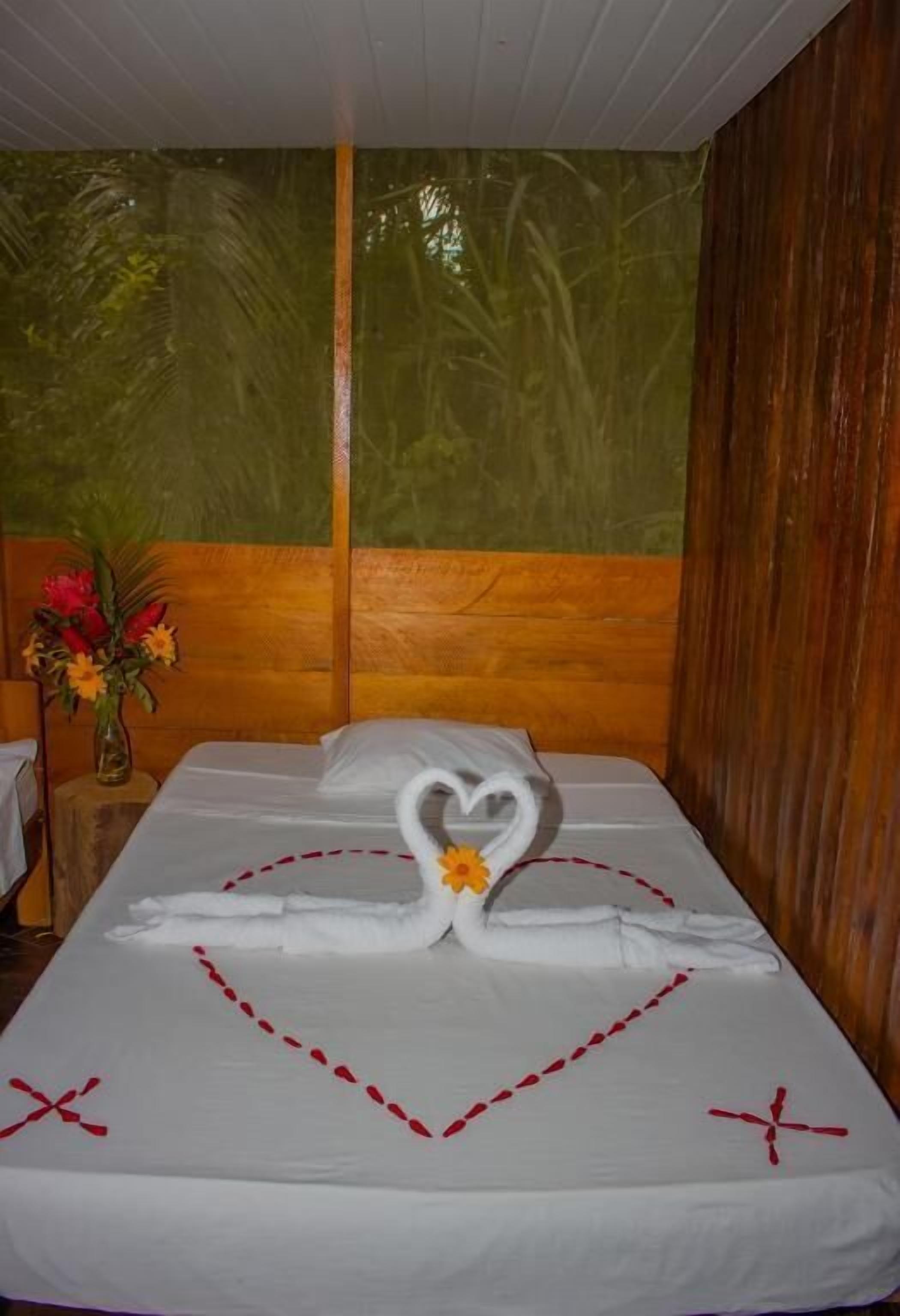 Een keurig opgemaakt bed met een handdoekarrangement in de vorm van een hart en een zwanen ontwerp, een vaas met bloemen, en een houten hoofdbord.