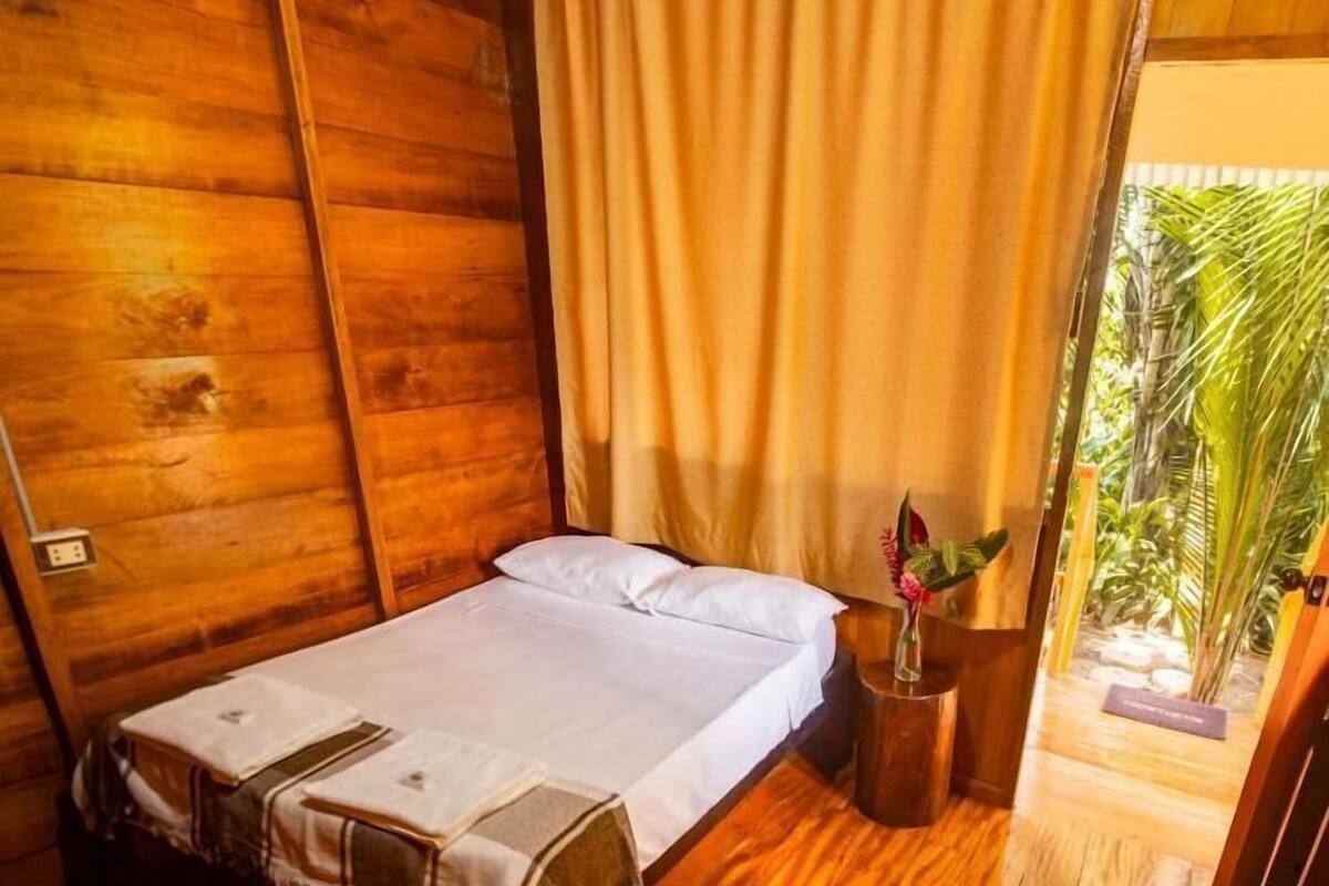 A single bed with a white bedspread and pillows, a wooden nightstand with a vase of flowers, and a wooden floor.