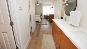Een hotelkamer met een houten vloer, een witte badkamer met een grote spiegel, een houten wastafel en een bed op de achtergrond.