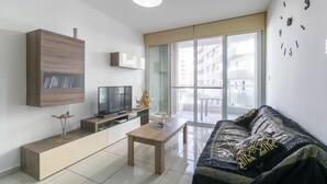 Una sala de estar moderna con televisor de pantalla plana, mobiliario de madera, puerta de vidrio que da a un balcón y un reloj de pared grande.