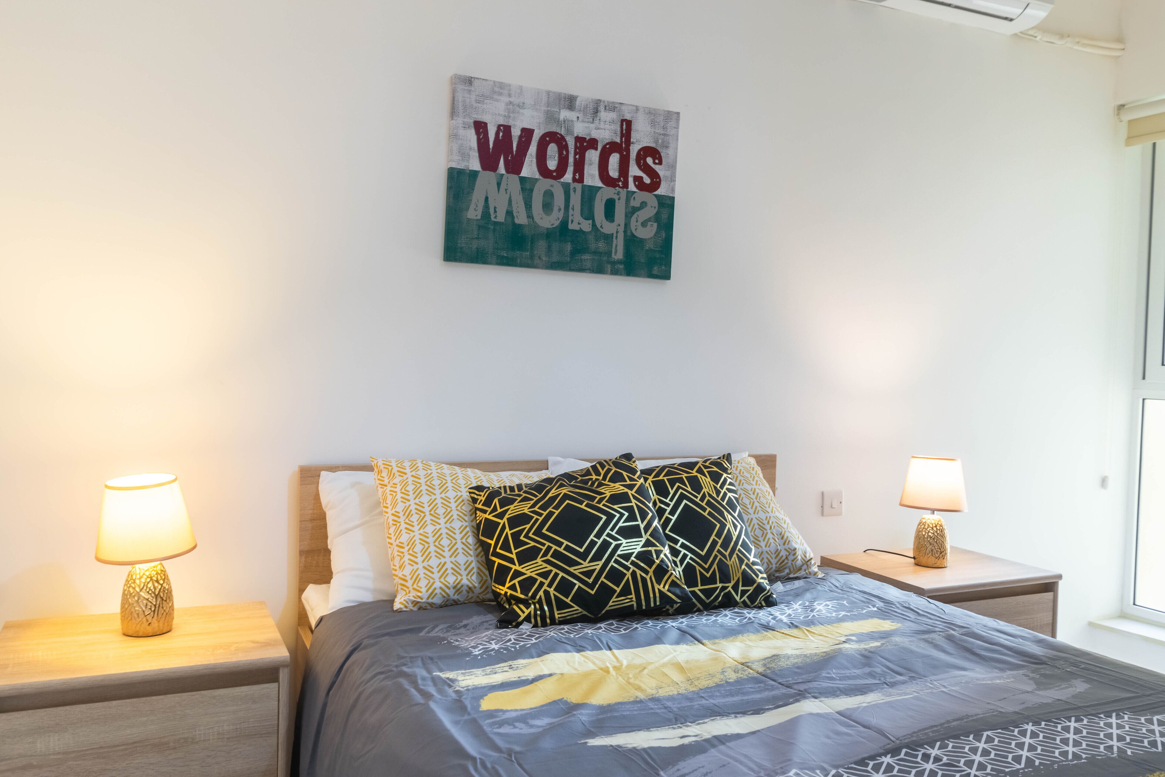 A neatly made bed with patterned bedding, two bedside lamps, and a wall-mounted sign.