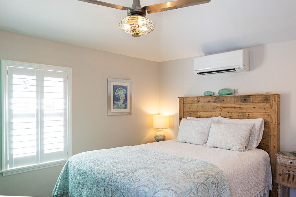 Ein Schlafzimmer mit einem hölzernen Kopfteil, einem Bett mit weißer Bettwäsche, einem Deckenventilator, einem Fenster mit Jalousien und einer Wand-Klimaanlage.