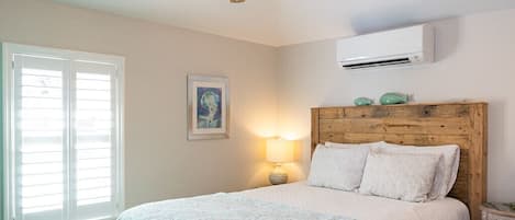 Ein Schlafzimmer mit einem hölzernen Kopfteil, einem Bett mit weißer Bettwäsche, einem Deckenventilator, einem Fenster mit Jalousien und einer Wand-Klimaanlage.