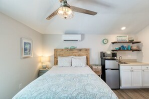 Une chambre avec une tête de lit en bois, un lit recouvert d’une couvre-lit à motifs, une table de chevet avec une lampe, un ventilateur de plafond, une kitchenette équipée d’un micro-ondes et d’un réfrigérateur, et une petite étagère avec des objets décoratifs.