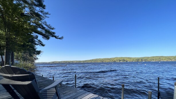 A wooden dock with a chair, overlooking a calm lake with boats and a forested shoreline.