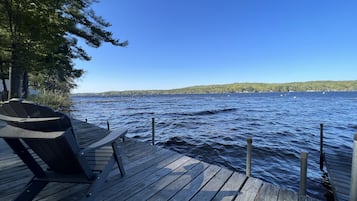 En träbrygga med en stol, med utsikt över en lugn sjö med båtar och en skogbeklädd strand.