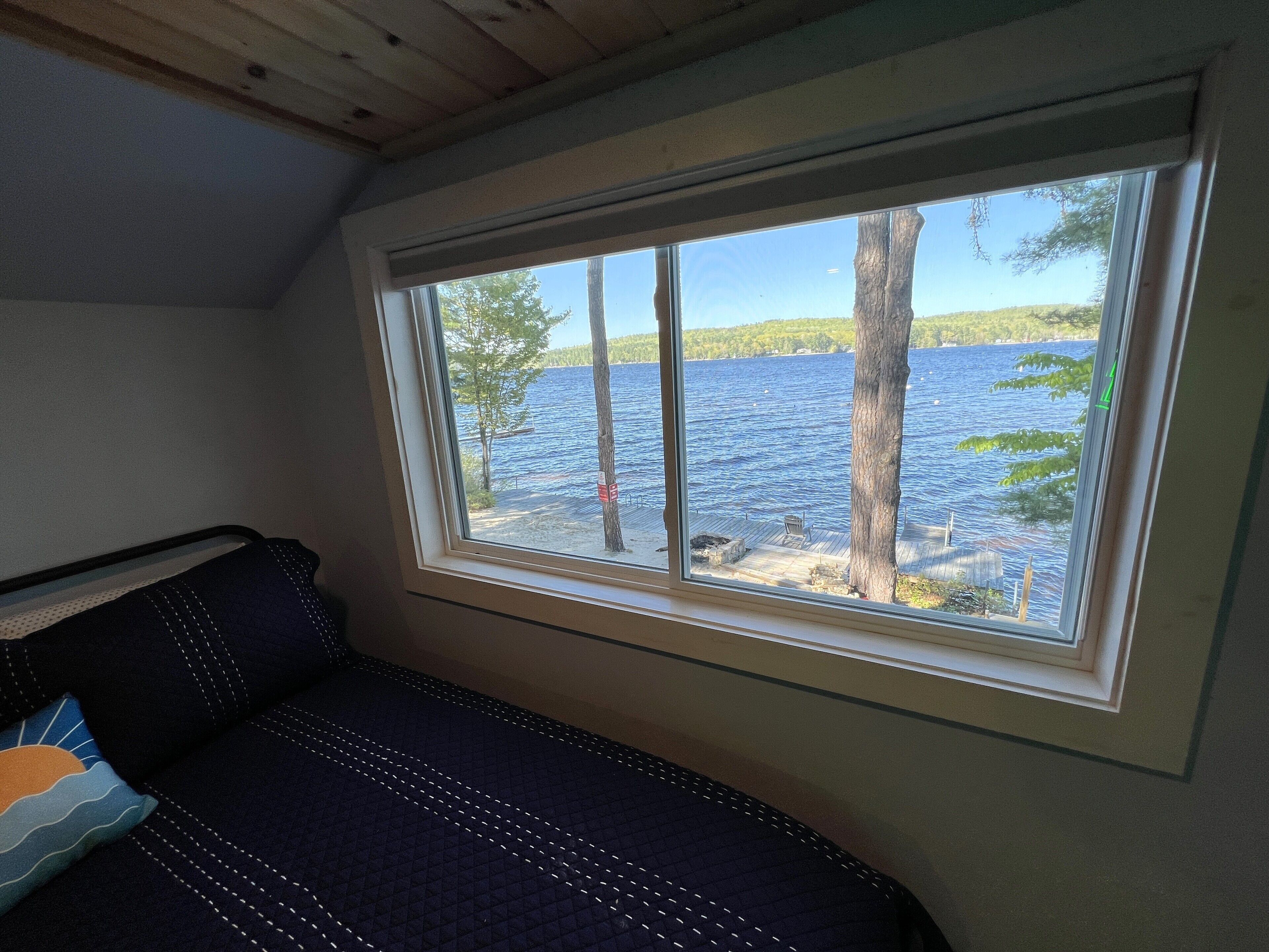 Una habitación con un ventanal grande que da a un lago, una cama con cubrecama acolchado y un techo de madera.