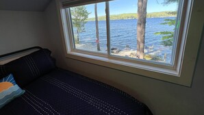 A room with a large window overlooking a lake, a bed with a quilted cover, and a wooden ceiling.