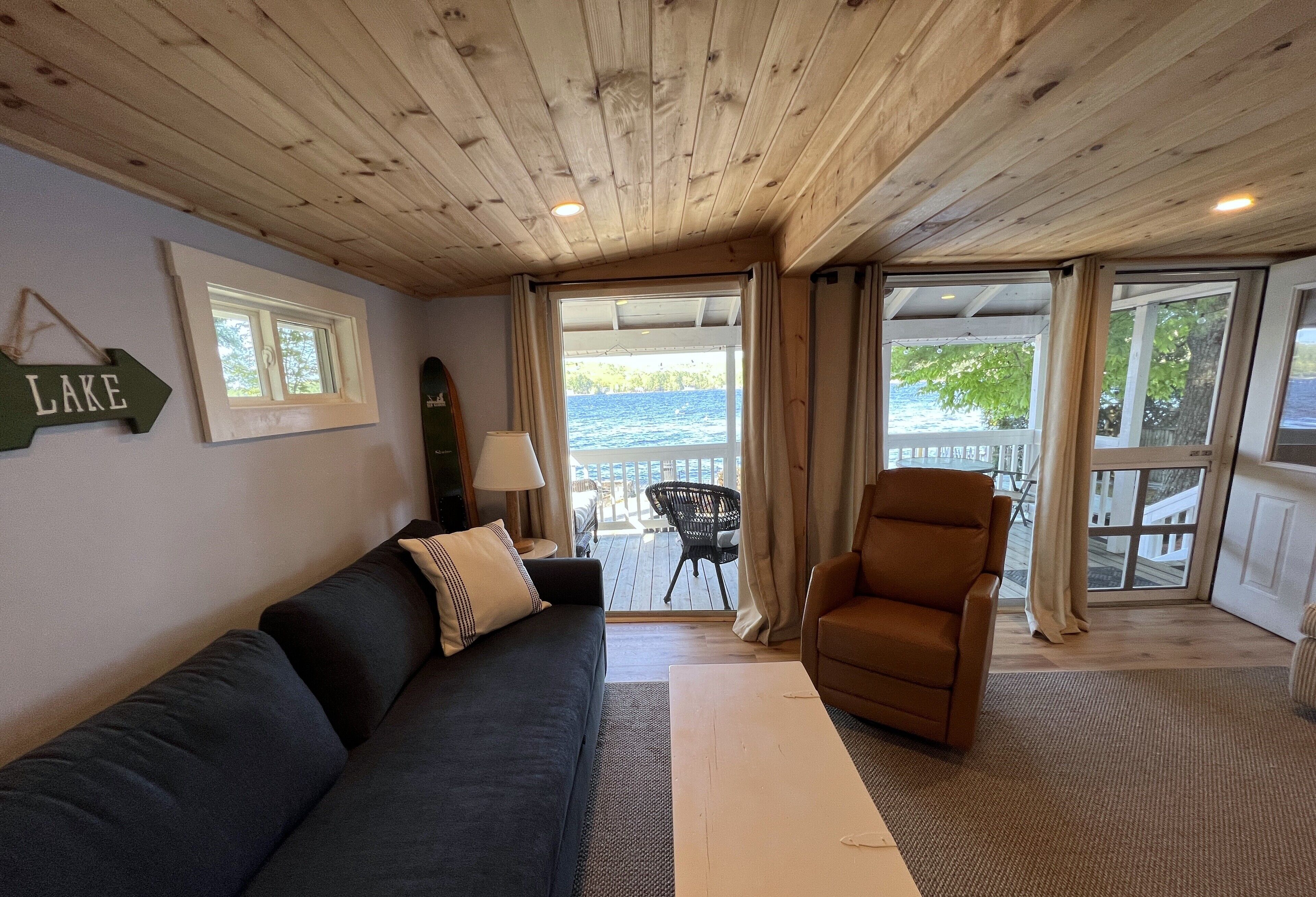 Un cuarto acogedor con un sofá azul oscuro, un sillón marrón y techo de madera. Se ve una ventana con vista al lago, y un cartel en la pared dice “LAKE”.