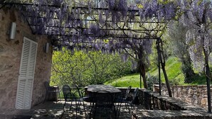 En udendørs terrasse med et bord og stole, en pergola dækket af lilla blomster, samt en stenvæg.
