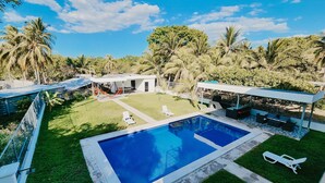 A modern outdoor pool area with lounge chairs, a barbecue, and a covered seating area, surrounded by lush greenery and palm trees. - Luxurious Villa with expansive room for family, couples/groups. 5 min beach walk (San Luis La Herradura)