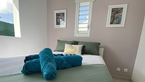 En snyggt bäddad säng med ett grönt överkast och flera kuddar, inklusive en grön och en vit. På väggen hänger två inramade konstverk, ett ovanför fönstret och ett till höger.