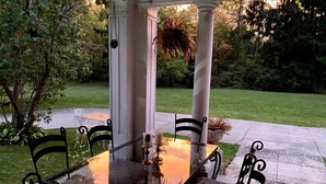 Une terrasse couverte avec une table en verre et des chaises en métal, entourée d’un jardin et d’un sentier.