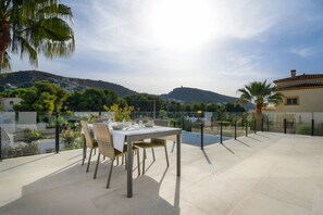 An outdoor patio with a dining table and chairs, a swimming pool, and a view of a hillside with buildings. - Graham Holiday Rentals - Magnolia (Moraira)