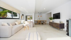 A modern living room with a sofa, ottoman, dining area, and a TV mounted on the wall. - Graham Holiday Rentals - Magnolia (Moraira)