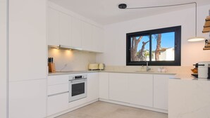 A modern kitchen with white cabinetry, a built-in oven, and a window with a view of trees. - Graham Holiday Rentals - Magnolia (Moraira)
