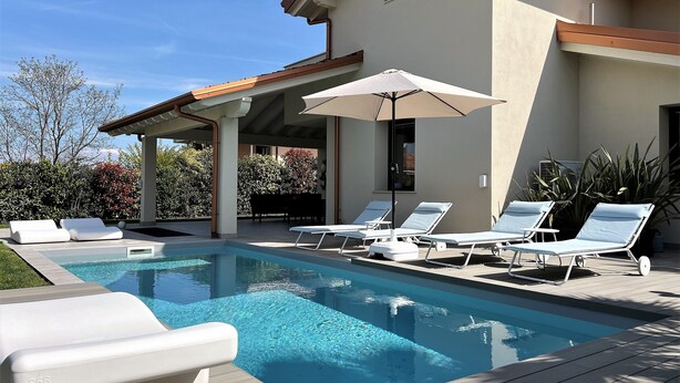 A swimming pool with lounge chairs and an umbrella, surrounded by a patio area with a house in the background. - Vacation home "Casa sulle colline di Barcuzzi" Pool, garden & barbecue (Barcuzzi)