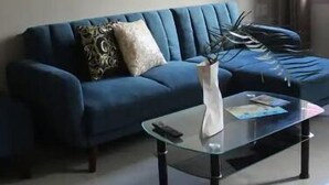Una sala de estar con un sofá azul, una mesa de centro de vidrio y una ventana con cortinas.
