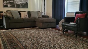 A living room with a sectional sofa, ottoman, patterned rug, a floor lamp, a black chair, and a window with curtains.