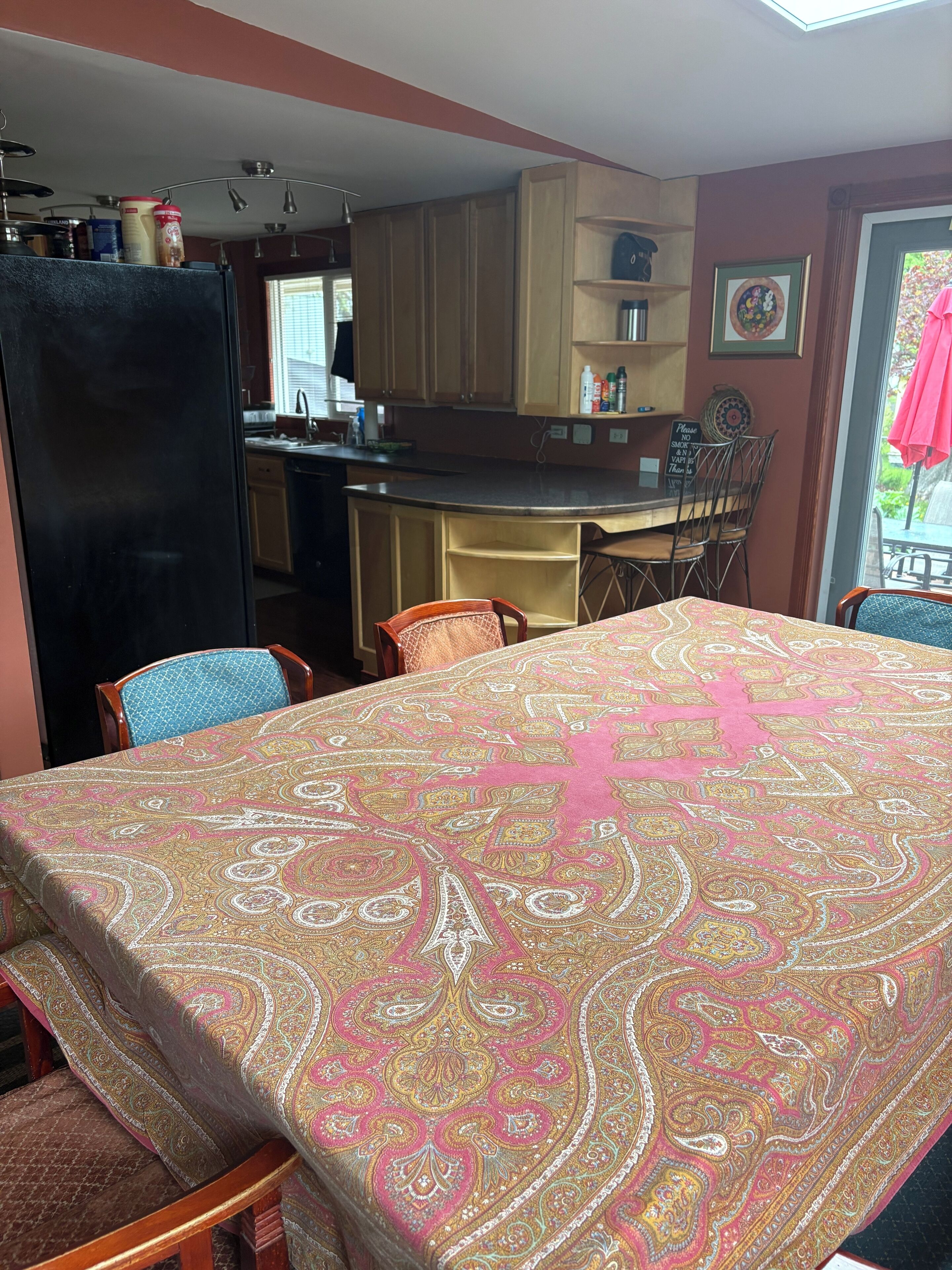 A dining area with a table covered in a patterned tablecloth, wooden chairs, and a kitchen area with cabinets and a refrigerator.