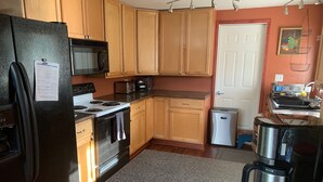 A kitchen with wooden cabinets, a black refrigerator, a microwave, a stove, a trash bin, and a coffee maker. - Spacious Home in Skagway Alaska (Skagway)