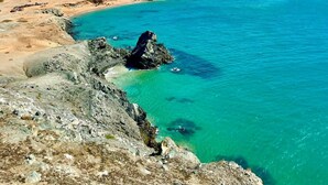 Un paysage côtier caractérisé par une eau turquoise et limpide, un terrain rocheux et de vastes plages de sable à l’horizon, le tout sous un ciel bleu parsemé de nuages.