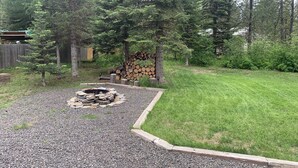 Un jardin avec un foyer entouré de gravier, une pile de bois de chauffage et une terrasse en bois.