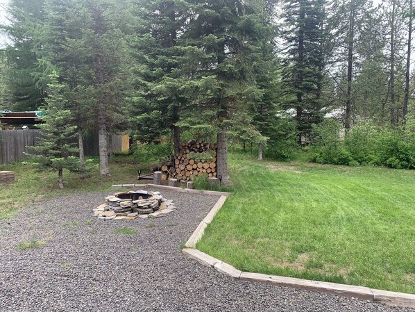 A backyard with a fire pit surrounded by gravel, a stack of firewood, and a wooden deck.