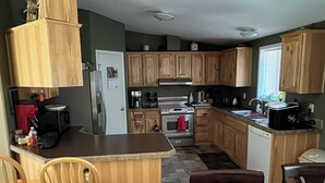 Une cuisine avec des meubles de rangement en bois, un réfrigérateur, une cuisinière et un four à micro-ondes. On y trouve deux chaises en bois et une table.