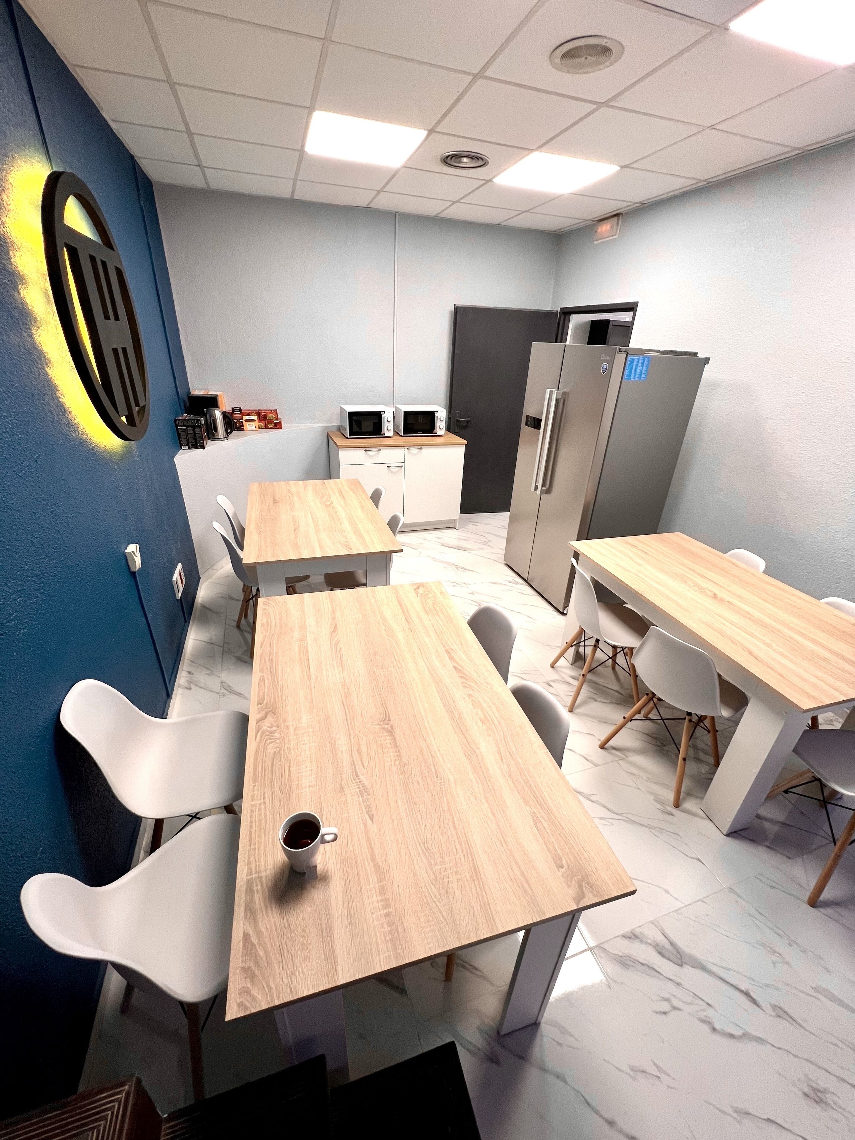 A modern kitchen with white cabinets, a microwave, and a refrigerator. There are wooden tables and white chairs.