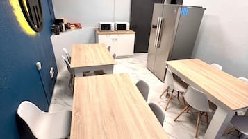 Una cocina moderna con armarios blancos, un microondas y un refrigerador. Hay mesas de madera y sillas blancas.