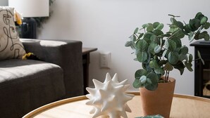 Olohuoneessa on pyöreä puinen pöytä, ruukkukasvi, valkoinen koriste-esine, seinällä oleva kehystetty kuva sekä harmaa sohva, jossa on lamppu.