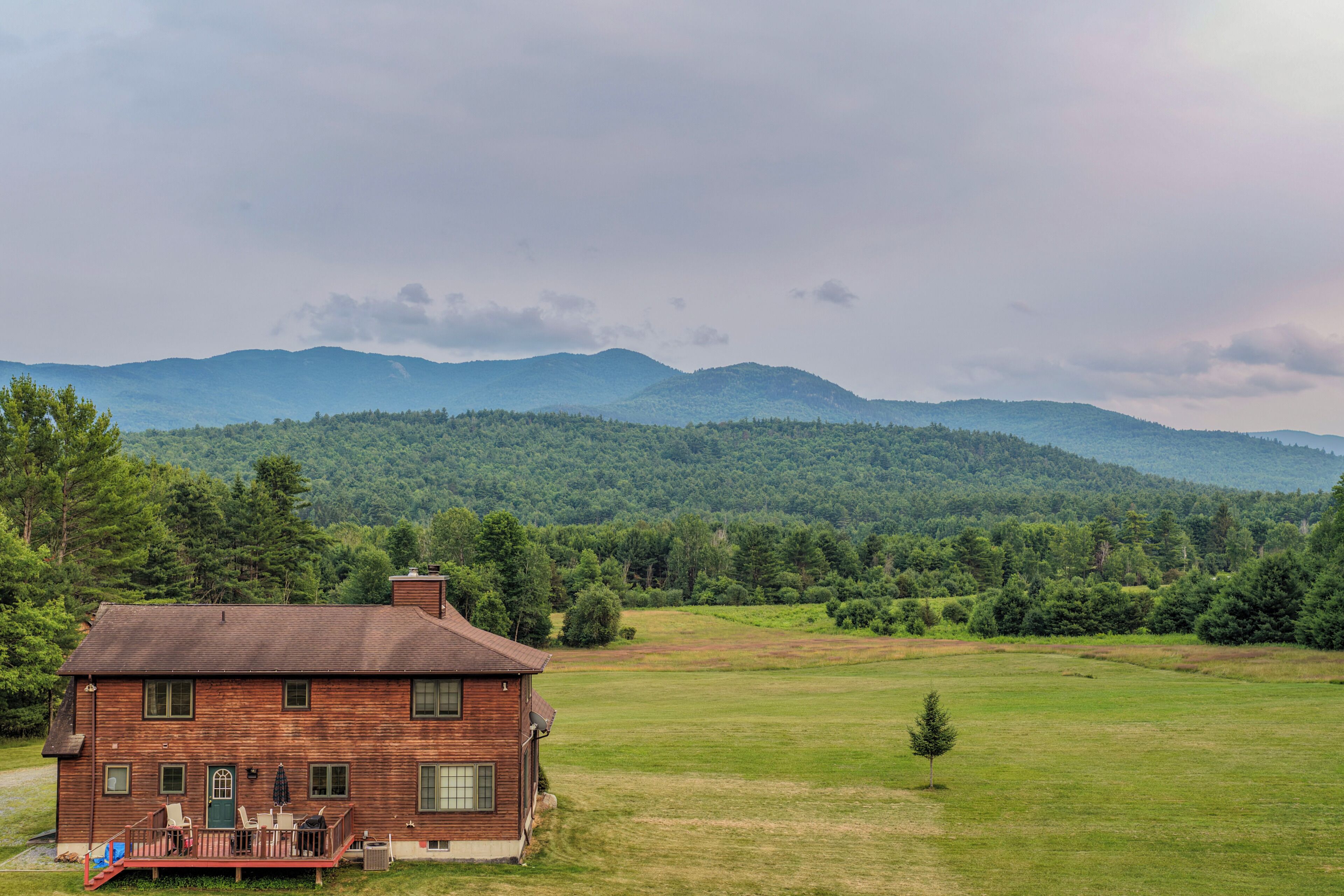 A wooden cabin with a deck, surrounded by a grassy field and forested hills.