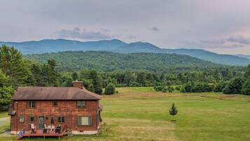 A wooden cabin with a deck, surrounded by a grassy field and forested hills.