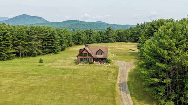 A two-story wooden house with a sloped roof, surrounded by a large grassy field and dense forest. - Deer Haven Lodge on 16 acres with mountain views and fireplace (Jay)