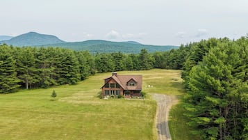 A two-story wooden house with a sloped roof, surrounded by a large grassy field and dense forest.
