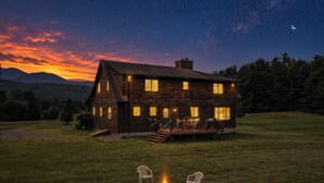 Ein Holzhaus mit Terrasse und Sitzgelegenheiten, umgeben von einer grünen Wiese und einem sternenübersäten Nachthimmel.