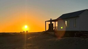 Una casa de playa con un ventanal grande y un balcón, ubicada en una playa de arena con el sol poniéndose al fondo.