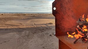 Una estructura metálica con un hueco, un fuego ardiendo a la derecha y una playa con arena y huellas en el fondo.