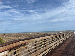 A wooden boardwalk with a metal railing, leading to a beach with dry grass and dunes.