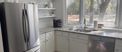 A modern kitchen with stainless steel appliances, white cabinetry, a granite countertop, and a window with blinds.