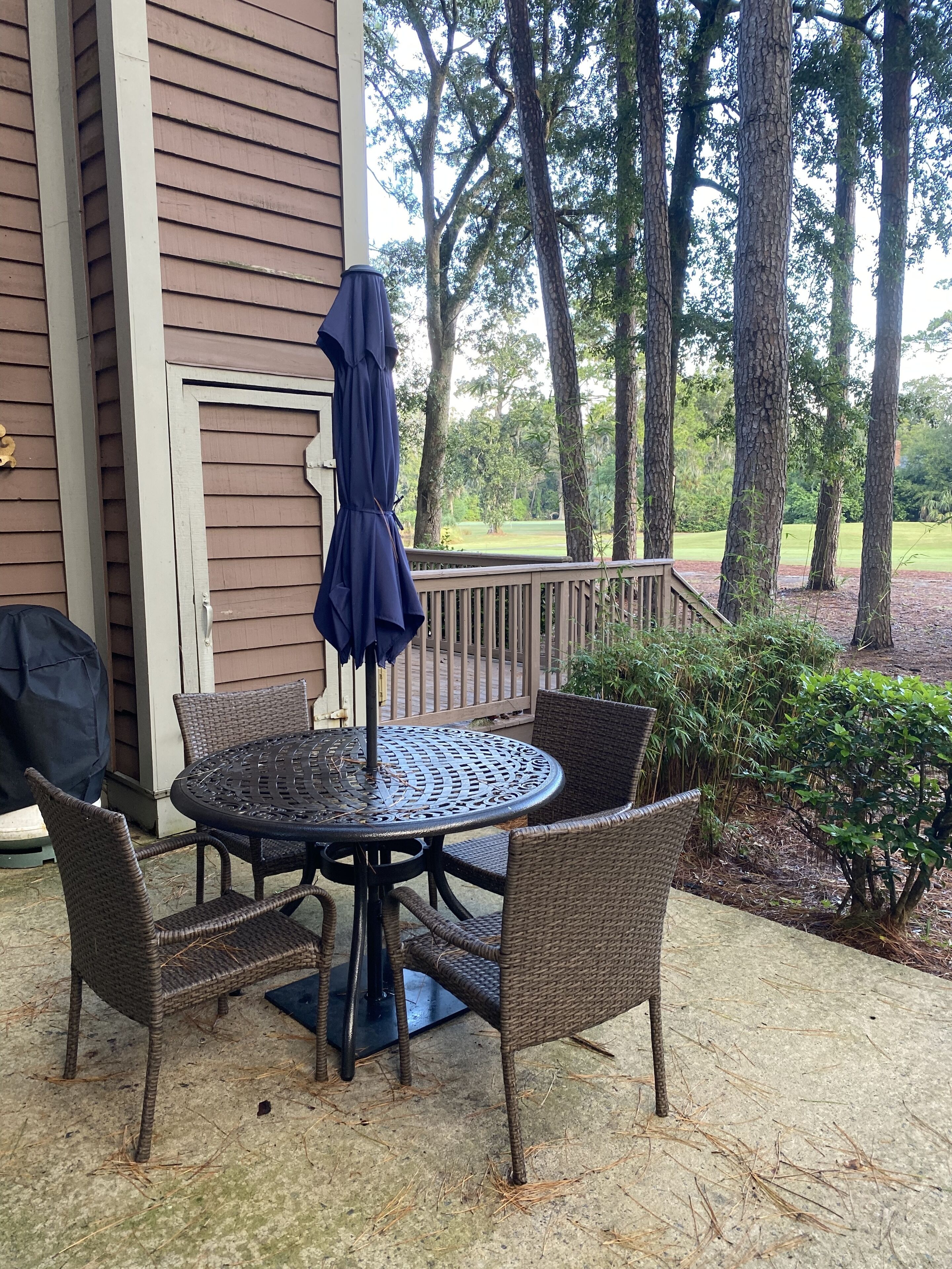 Een terras met een ronde tafel, vier rieten stoelen, een blauwe parasol en een barbecue.
